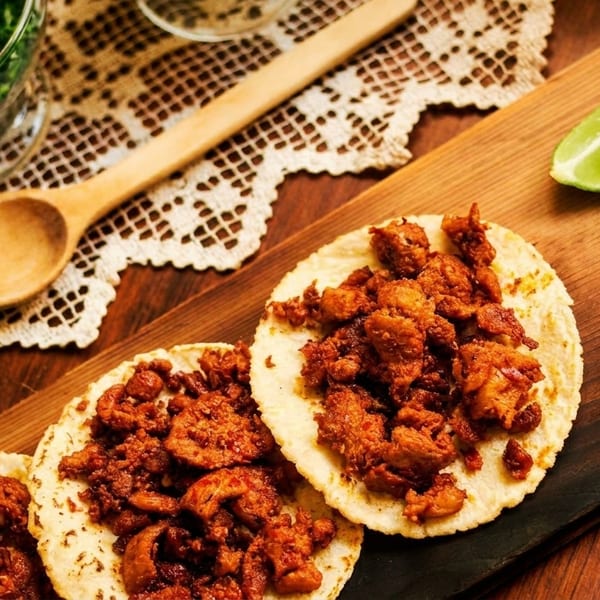 Three tacos with seasoned meat, lime wedges, dried chilies, and cilantro on a wooden board.