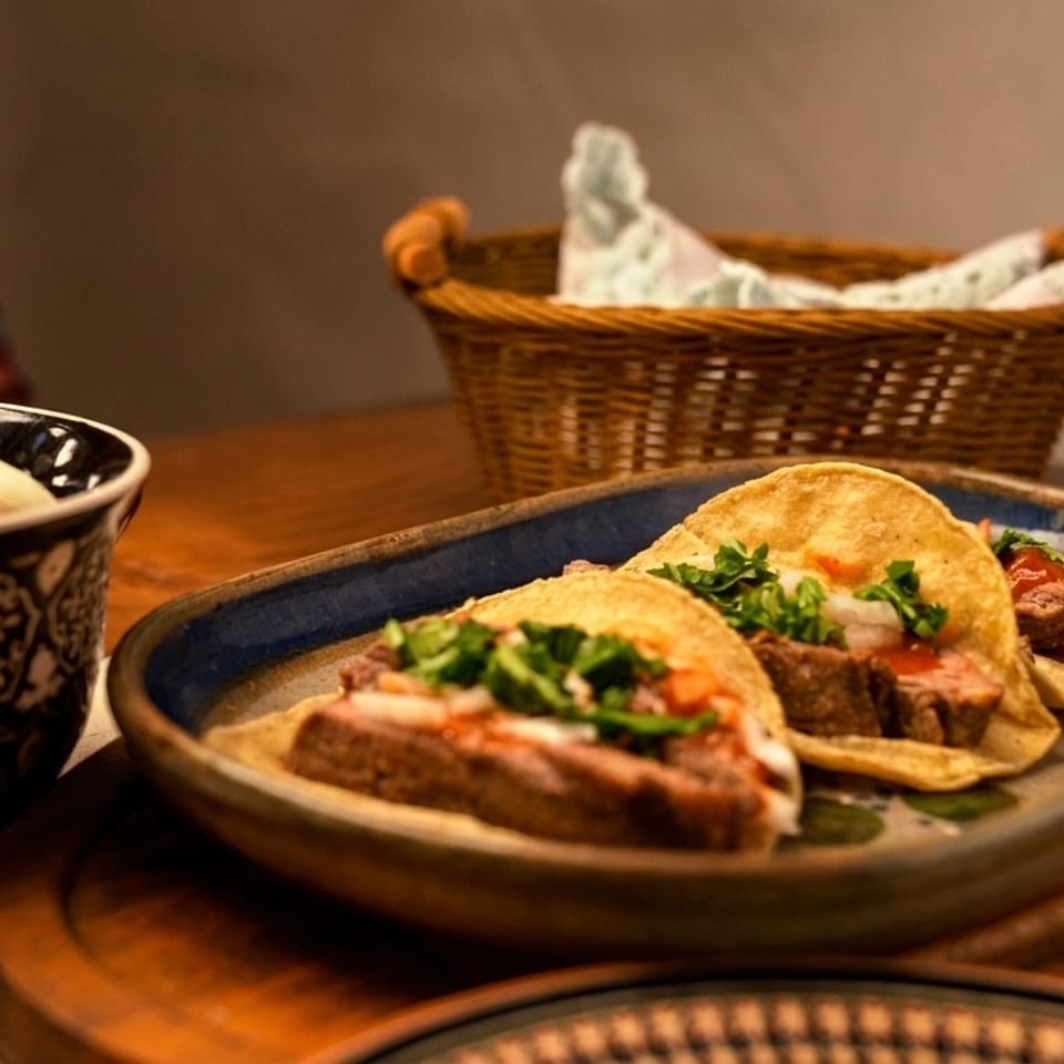 Three beef tacos with cilantro and onions on a blue plate, with a basket and bowl in the background.